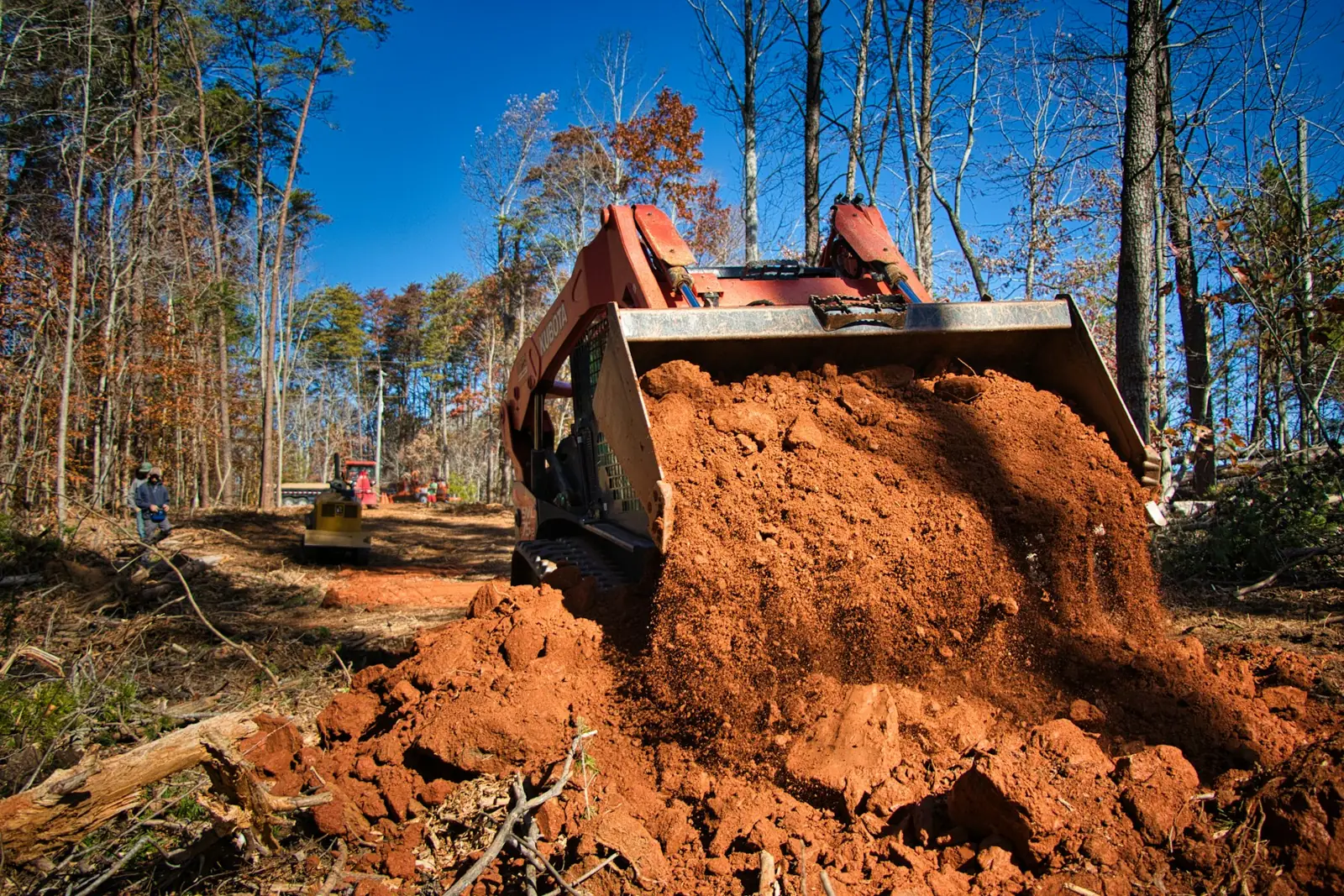 Compact track loader pushing red clay in a wooded clearing
